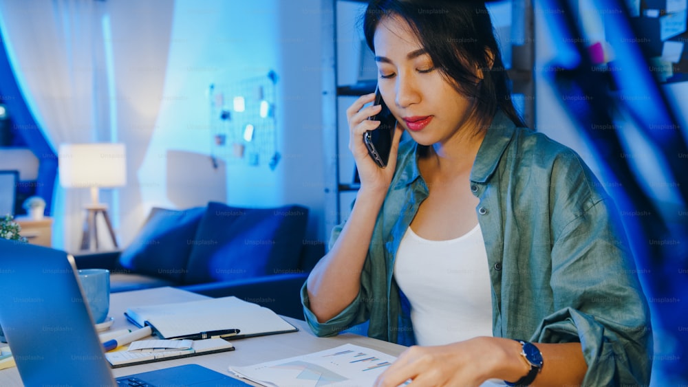 Mujeres asiáticas independientes que usan la computadora portátil hablan por teléfono empresarias ocupadas que trabajan a distancia en la sala de estar. Sobrecarga de trabajo desde casa por la noche, trabajo remoto, distanciamiento social, cuarentena por coronavirus.