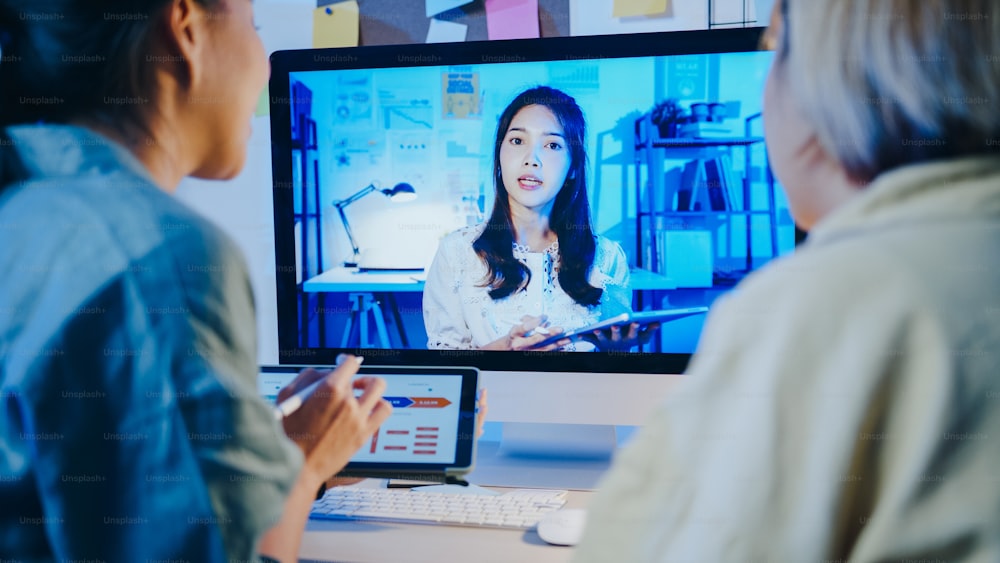 Las empresarias asiáticas que usan computadoras de escritorio hablan con colegas sobre el plan en una reunión de videollamada en la sala de estar. Sobrecarga de trabajo desde casa por la noche, trabajo remoto, distanciamiento social, cuarentena por coronavirus.