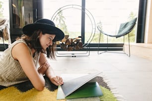 Beautiful stylish woman relaxing with book on cozy rug on background of fireplace in modern chalet. Young female in casual clothes reading magazine on floor. Leisure and vacation time