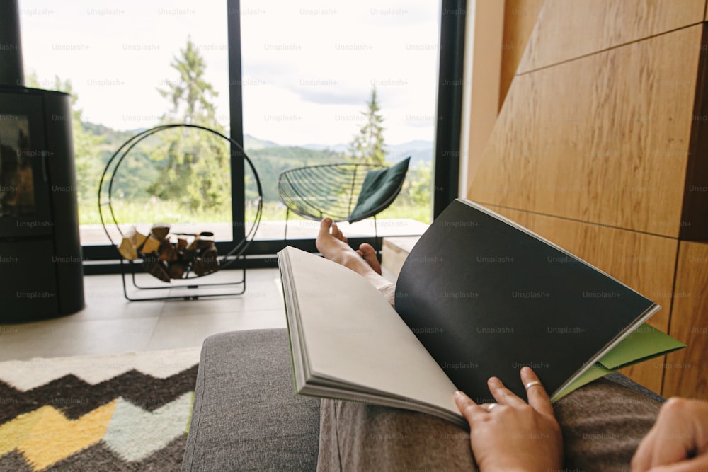 Schöne weibliche Füße und Buch auf dem Hintergrund des Kamins und des großen Fensters im modernen Chalet. Junge Frau barfuß entspannend mit Buch in komfortablem Zuhause, gemütlicher Urlaub. Freizeit