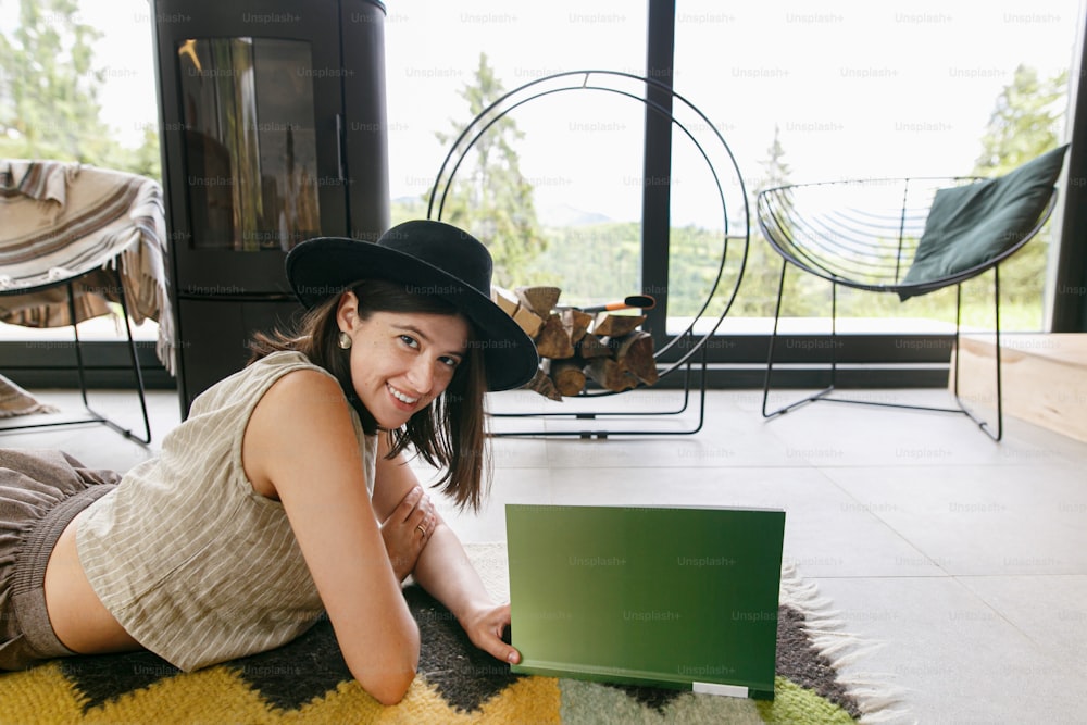 Beautiful stylish woman relaxing with book on cozy rug on background of fireplace in modern chalet. Young happy female in casual clothes reading magazine on floor. Leisure and vacation time