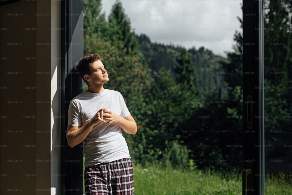 Handsome man enjoying hot coffee in sunny morning on background of mountain hills. Calm tranquil moment. Young man in pajamas relaxing with cup of coffee on porch. Space for text