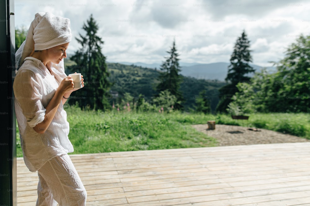 Beautiful young woman enjoying coffee in sunny morning on background of mountain hills. Calm tranquil moment. Beautiful female in white pajamas and towel after shower relaxing with cup of coffee