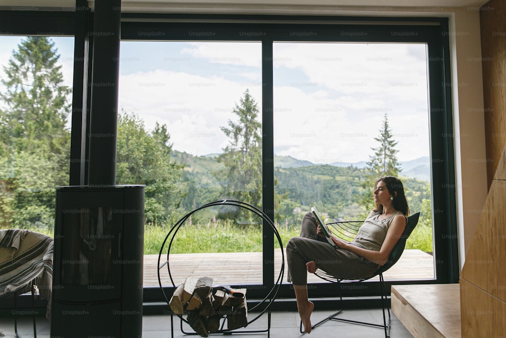 Beautiful stylish woman reading book on chair at fireplace with firewood on background of mountain hills. Young female in casual clothes relaxing in modern chalet with amazing view from window