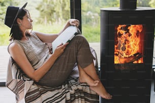 Beautiful stylish woman reading book on chair at warm burning fireplace on background of big window. Young female in casual clothes relaxing in modern chalet, cozy vacation. Leisure time