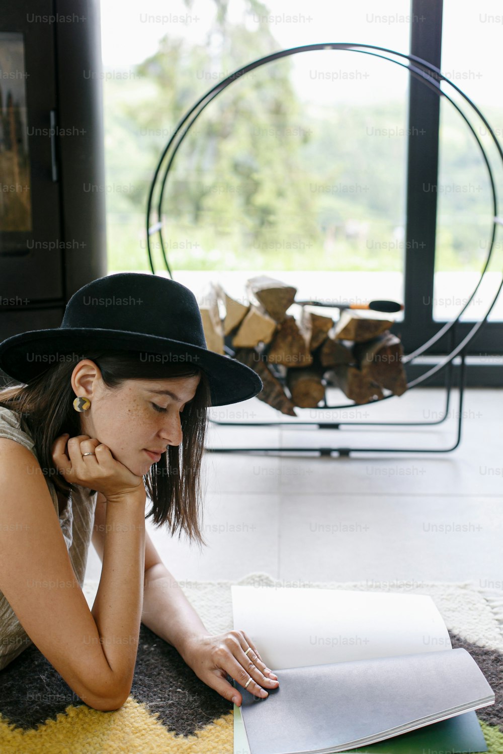 Beautiful stylish woman reading book on floor on background of fireplace and big window in modern chalet. Young female in casual clothes relaxing on cozy rug with magazine. Leisure and vacation time