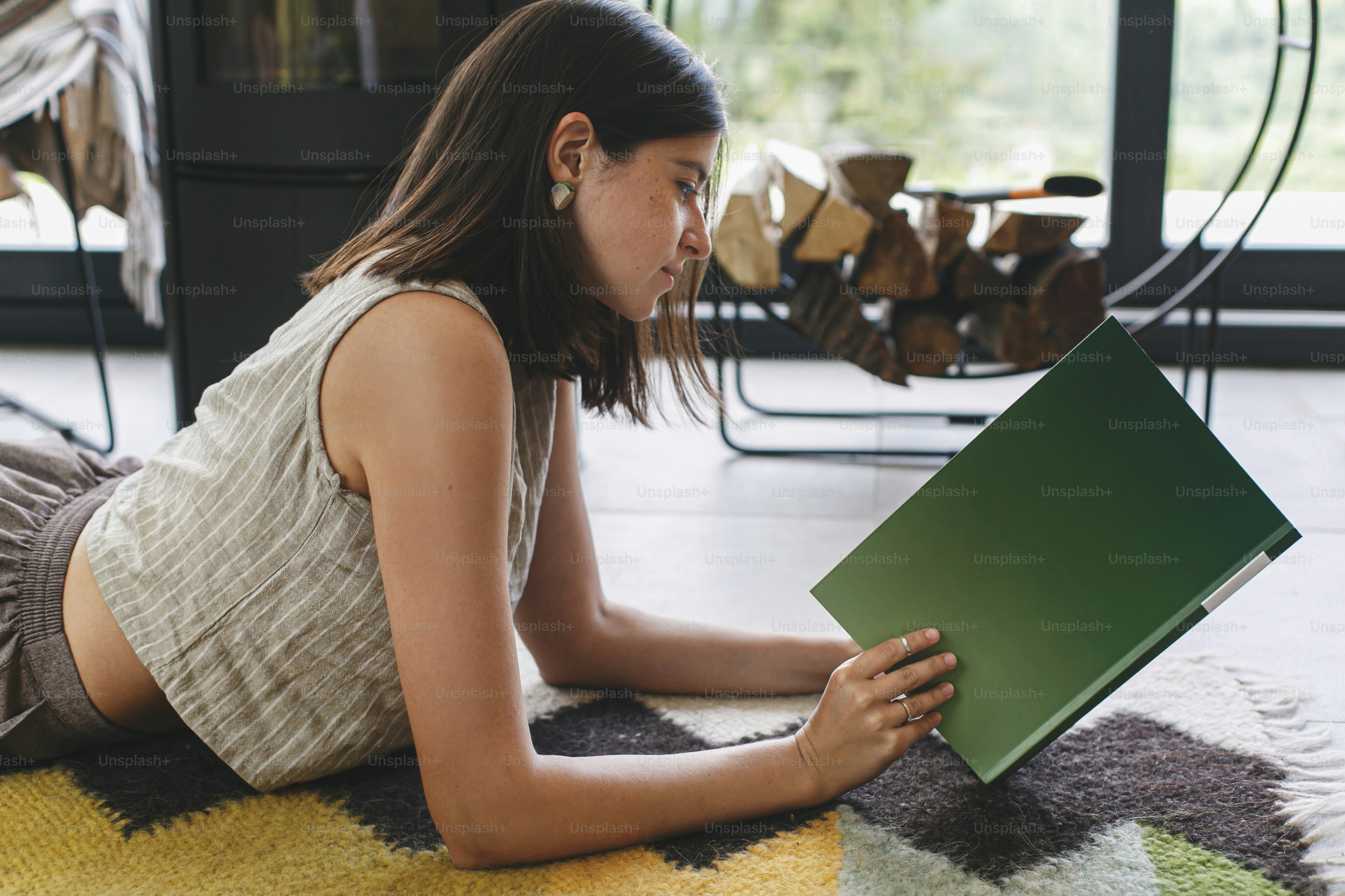 Beautiful stylish woman relaxing with book on cozy rug on background of fireplace in modern chalet. Young female in casual clothes reading magazine on floor. Leisure and vacation time