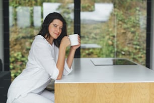 Beautiful happy woman enjoying hot coffee in sunny morning  in modern kitchen in chalet in mountains. Calm carefree moment. Young female in white pajamas relaxing with cup of coffee