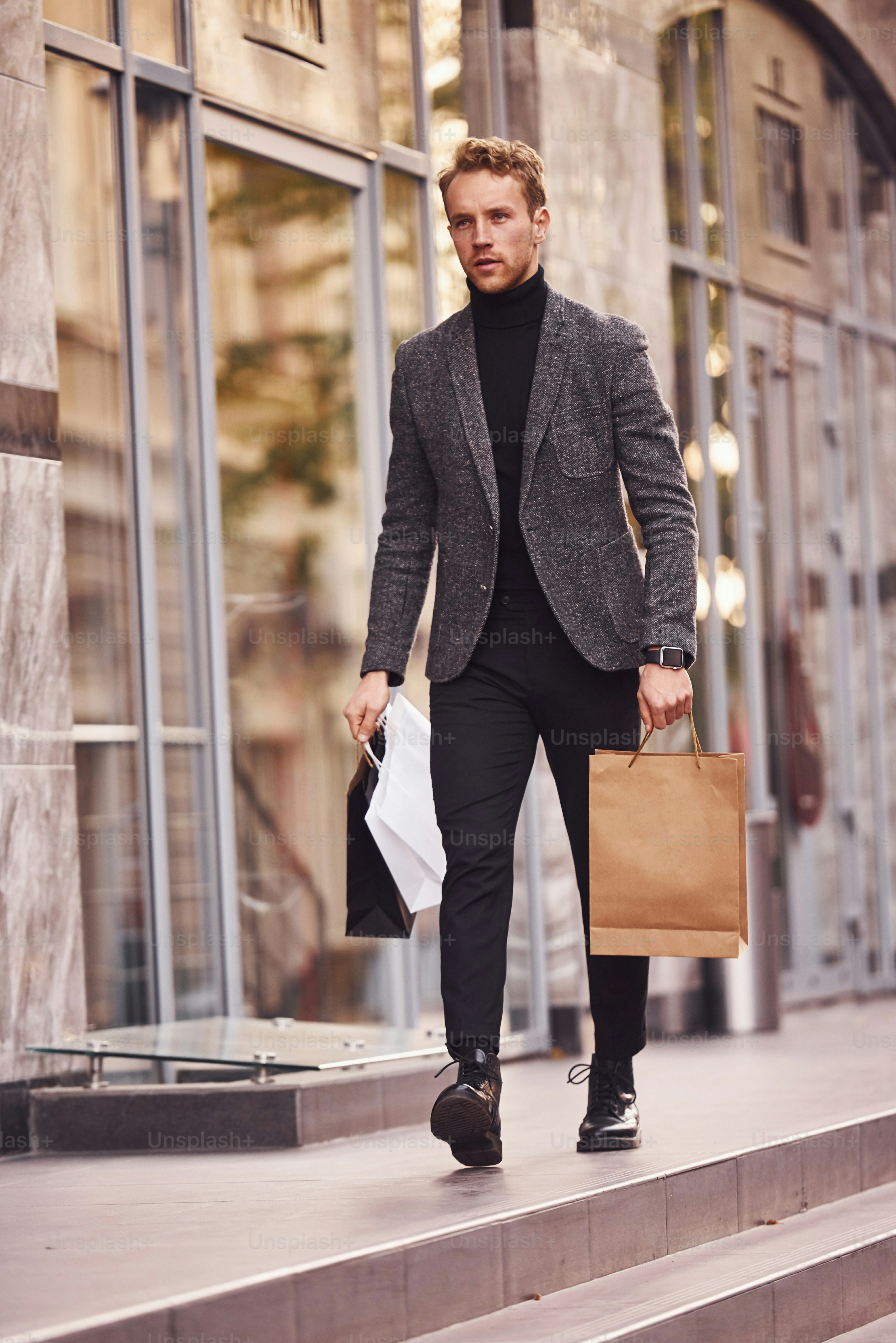 Man in elegant formal wear and with shopping packages is outside against modern building