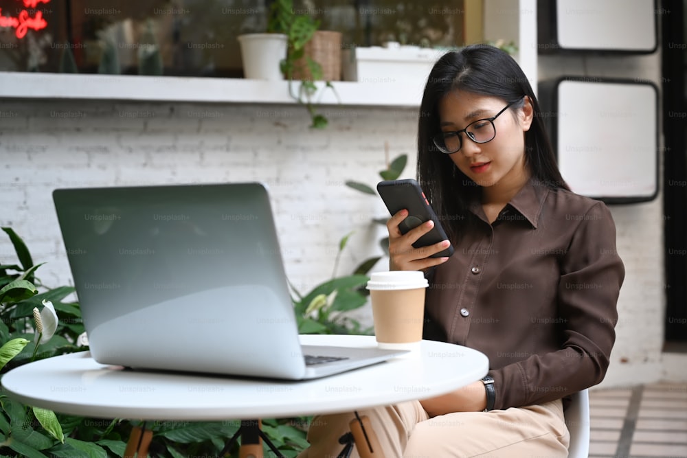 Mujer joven casual enviando mensajes de texto o masajeando en un teléfono inteligente mientras se sienta en un café al aire libre.