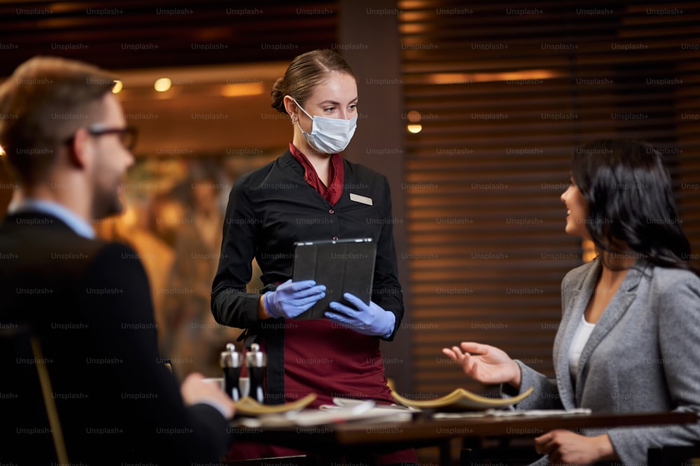 Hospitable waitress wearing mask and gloves taking food order from a lovely couple at a dinner table