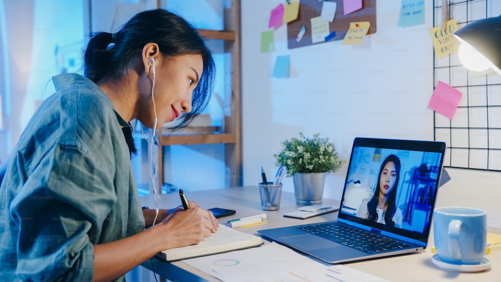 Asia businesswoman using laptop talk to colleagues about plan in video call meeting at living room. Working from house overload at night, remotely work, social distancing, quarantine for coronavirus.