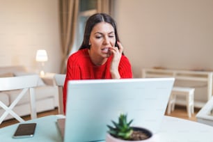 Nervous woman looking at laptop and biting her fingernails at home. Anxious depressed woman sitting with laptop and biting nails, looking nervous worried, scared of deadline stressful job.