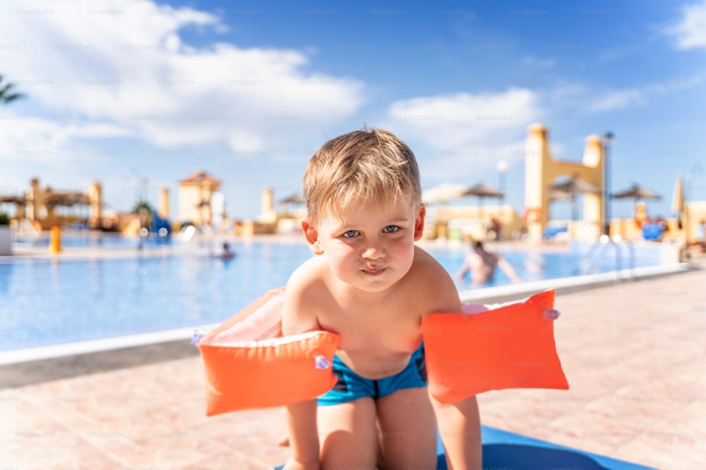 Kind mit aufblasbaren Armbinden in der Nähe des Schwimmbads. Kleiner Junge, der im Außenpool des tropischen Resorts schwimmen lernt. Sommerferien.