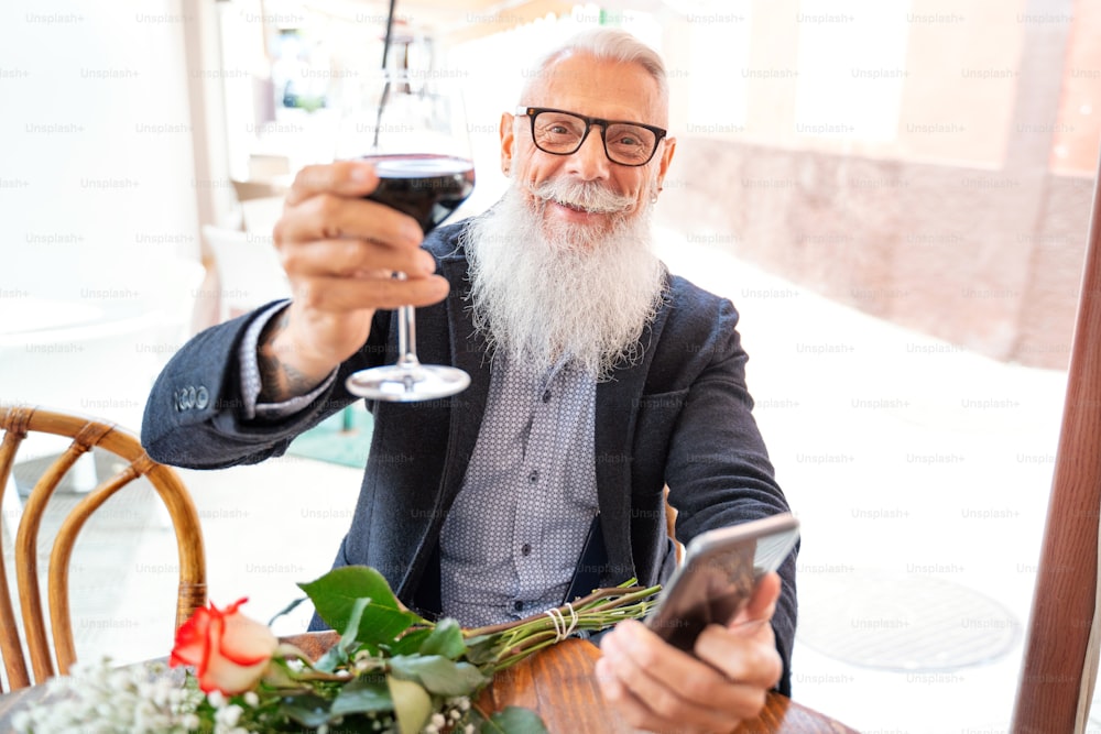 Senior man with white beard and mustache holding glass of wine, smiling to the camera, waiting for his date with flowers on the restaurant. Adult handsome men using mobile phone. Real people emotions. Happy lifestyle.
