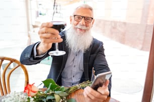白いあごひげを生やし、口ひげを生やした年配の男性がワイングラスを持ち、カメラに向かって微笑み、レストランで花を持ってデートを待っています。携帯電話を使う大人のハンサムな男性。実在の人物の感情。幸せなライフスタイル。