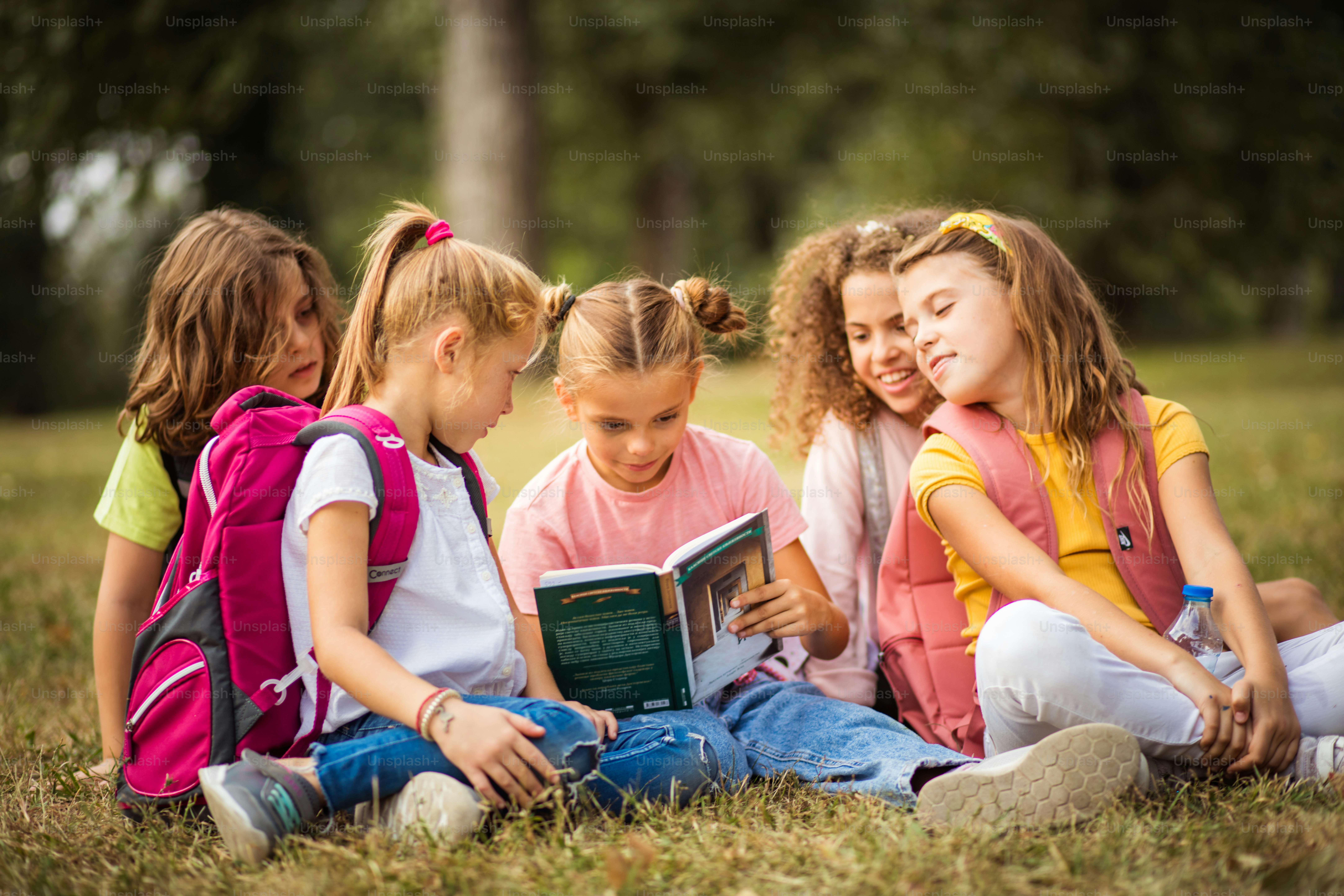 Large group of school kids having fun in nature. Little girl reading book to her friends.