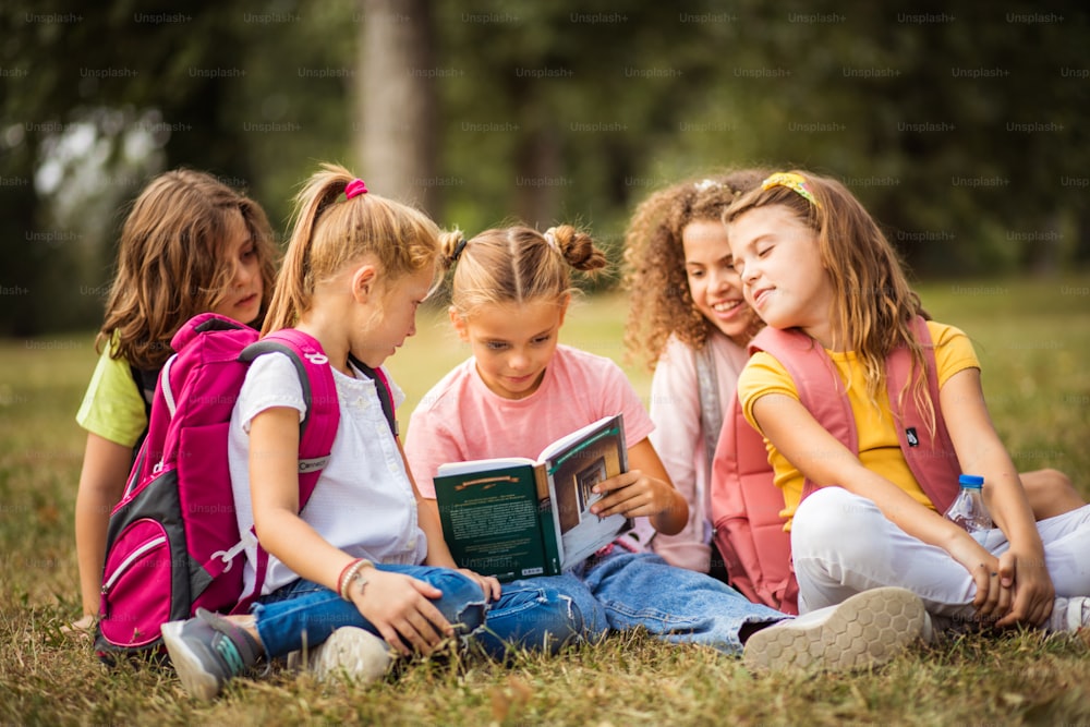 Large group of school kids having fun in nature. Little girl reading book to her friends.