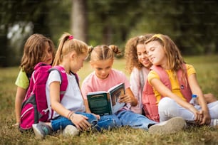 Large group of school kids having fun in nature. Little girl reading book to her friends.