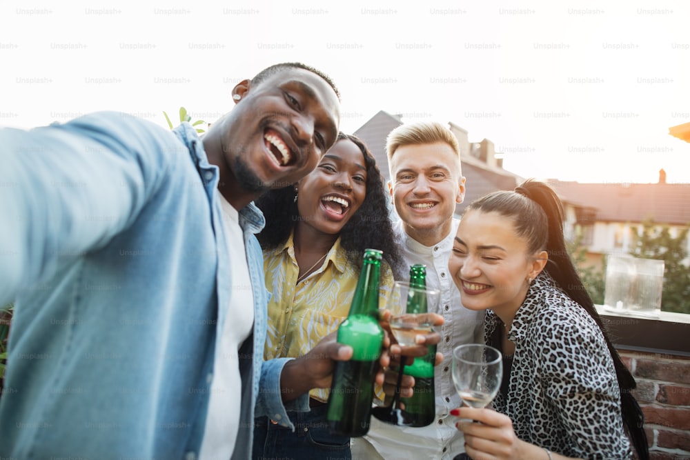 Gioiosi amici multiculturali in abiti eleganti che scattano selfie sullo smartphone con bevande alcoliche in mano. Concetto di persone, tecnologia e tempo di festa.
