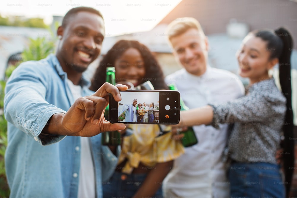 Quattro persone multiculturali in abiti eleganti che utilizzano uno smartphone moderno per scattare selfie durante la festa sul tetto. Giovani amici felici che sorridono, bevono, chiacchierano.