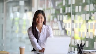 Happy young female employee sitting in modern office with laptop computer and smiling to camera.