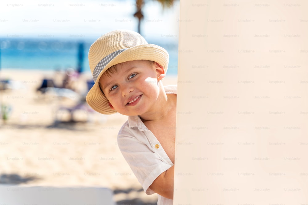 Niño sonriente con sombrero jugando en la playa tropical de arena. Vibraciones veraniegas. Mucho espacio de copia.