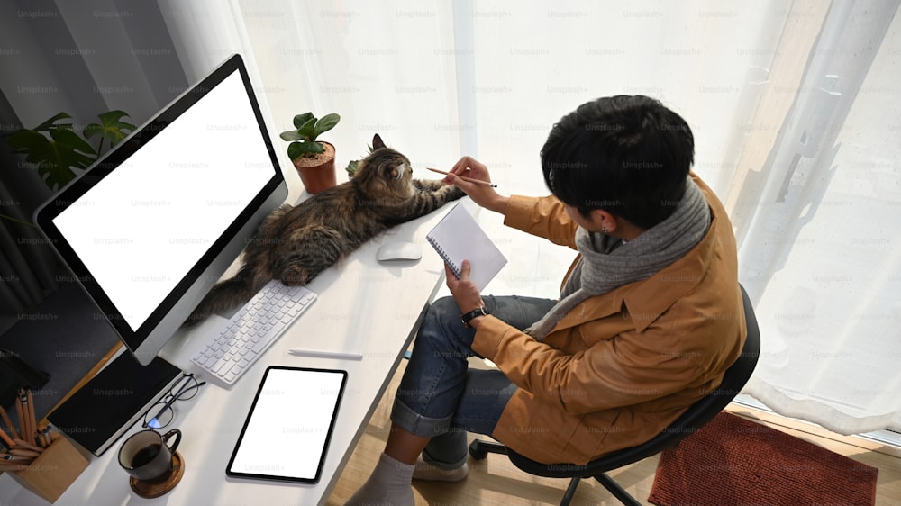 Vista acima do jovem sentado na frente do computador com seu gato e segurando o caderno.