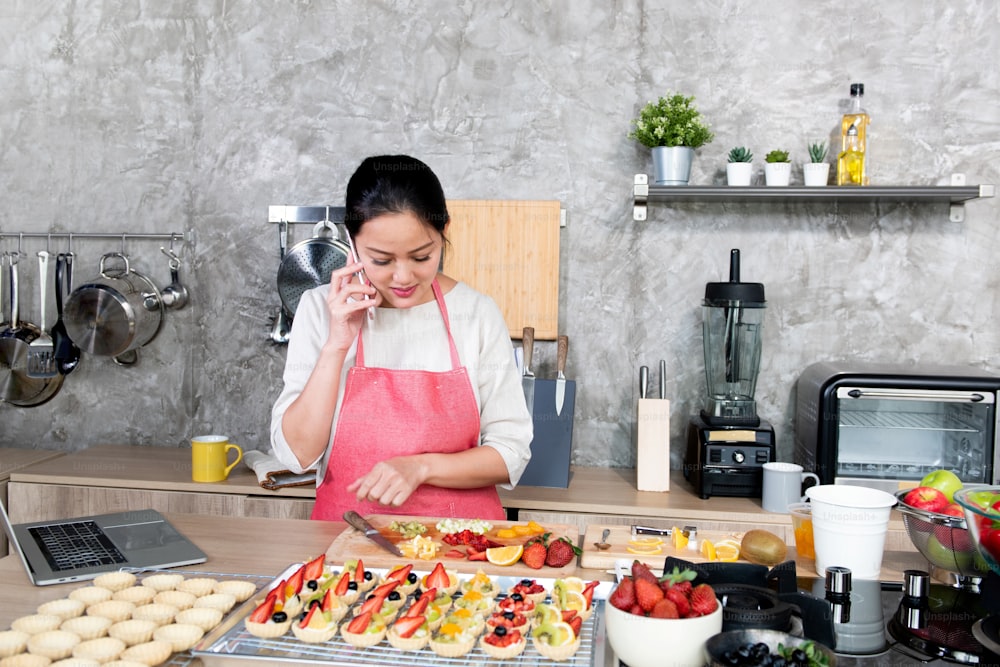 Asian woman making fruit tart for online selling delivery. Female small business bakery shop owner entrepreneur working in laptop computer and mobile phone get order for customer. Small business work at home concept.