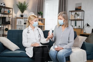 Professional family gp doctor in mask consulting elderly 60 aged woman patient at home. Female nurse showing a new oral spray to her mature patient, talking about correct use and dosage.