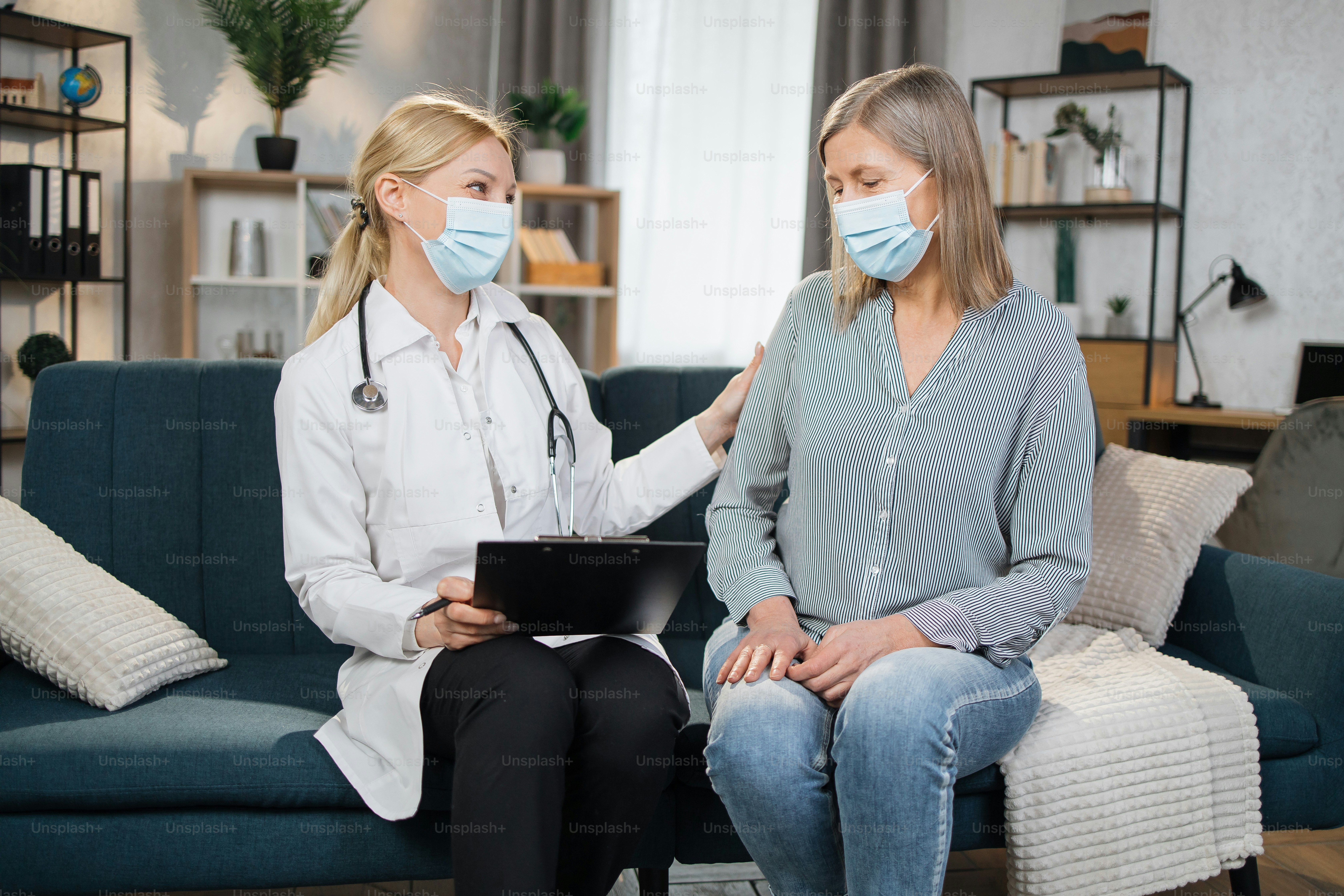 Happy blond female doctor consulting elderly woman at home, telling about good checkup result, holding clipboard with medical history or anamnesis