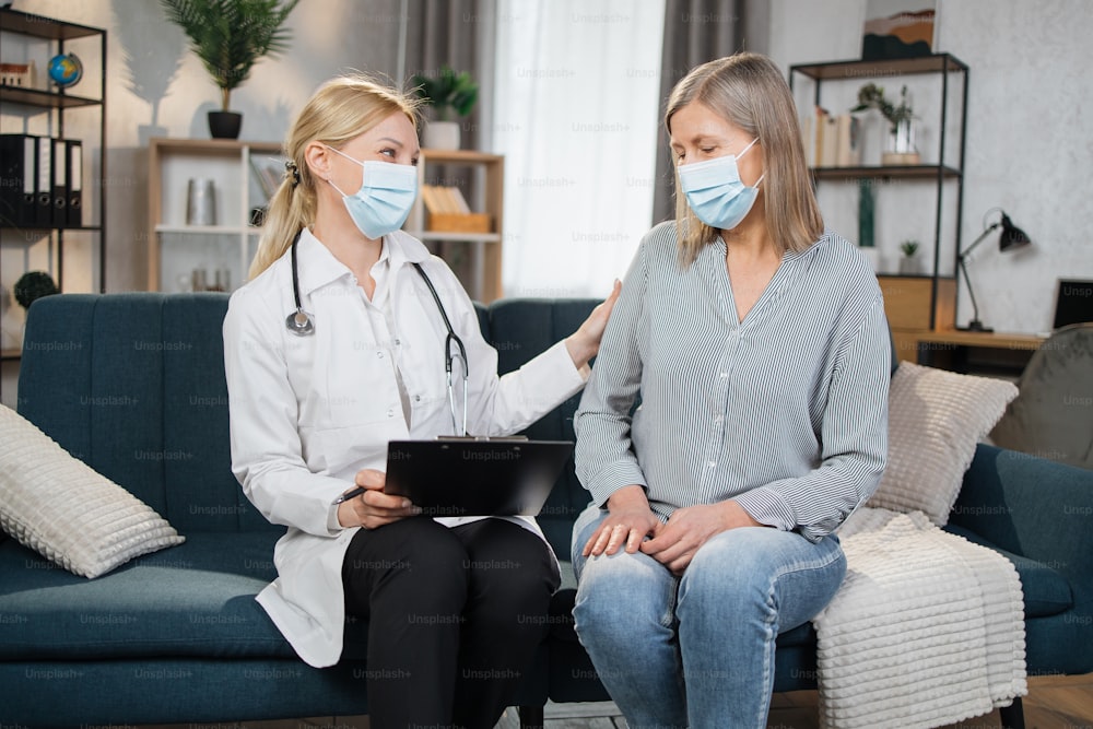 Happy blond female doctor consulting elderly woman at home, telling about good checkup result, holding clipboard with medical history or anamnesis