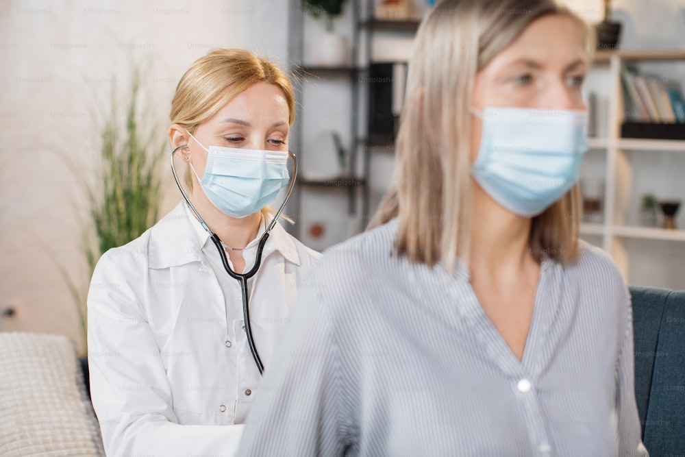 Close up de uma mulher médica caucasiana de jaleco branco e máscara facial protetora, auscultando sua paciente caucasiana sênior, usando um estetoscópio, durante a visita domiciliar. Tratamento contra gripe e covid-19