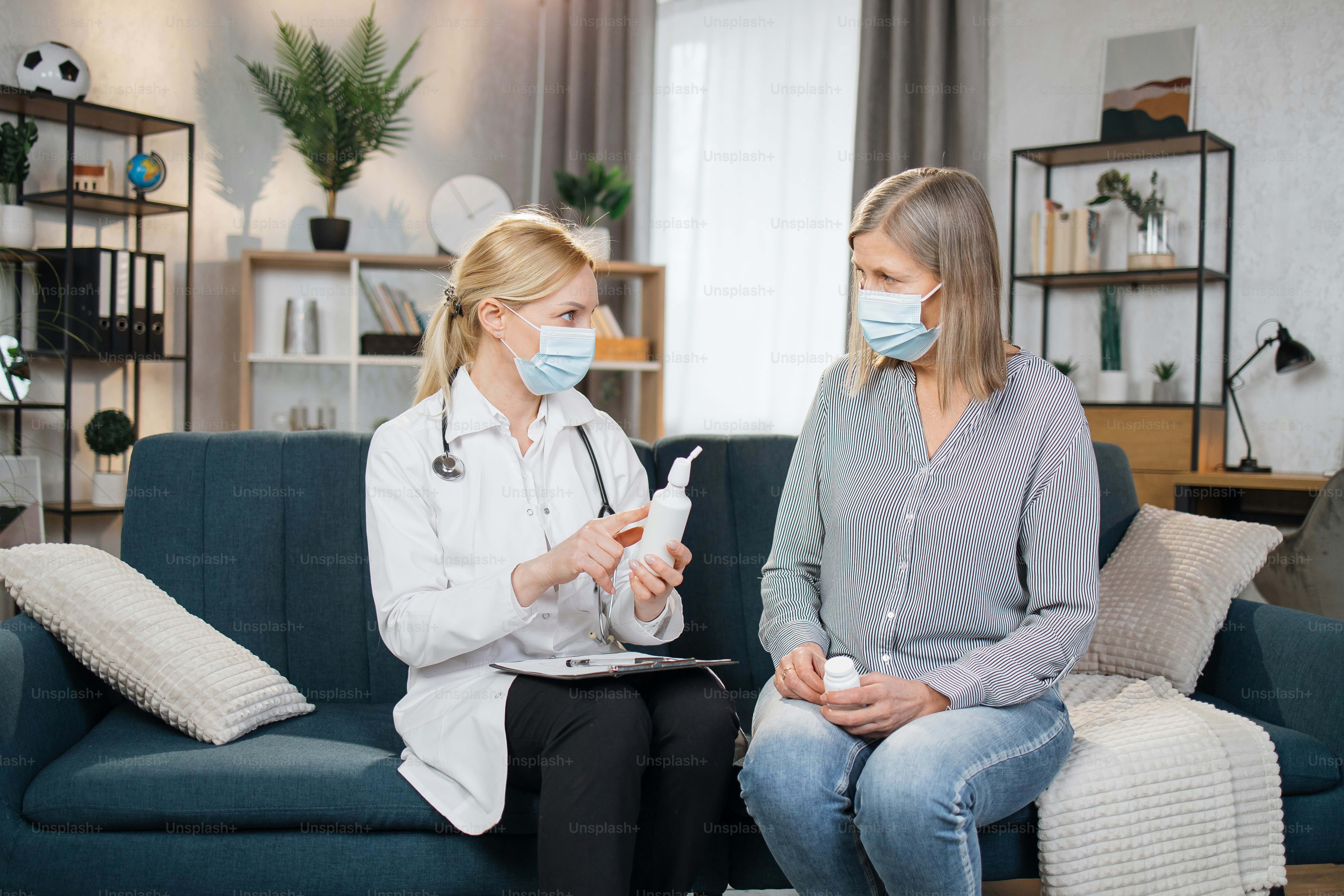 Serious disturbed senior woman in medical mask listening to female doctor visiting her at home, and talking about using of the spray for sore throat, dosage, indications and contraindications.