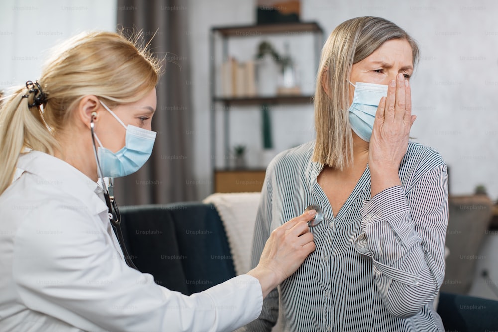 Home treatment of virus infection, coronavirus pandemic, Covid-19 outbreak. Female doctor during home visit examining sick senior woman patient in face mask, coughing, having chest pain.
