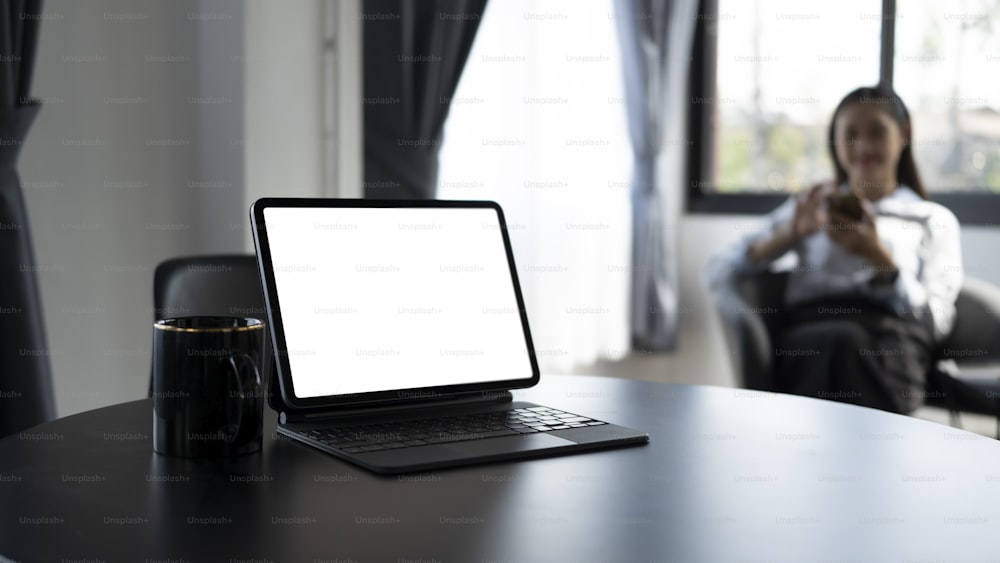 黒い木製のテーブルの上にコンピューターのタブレットとコーヒーカップのモックアップがあり、背景にビジネスウーマンが座っています。