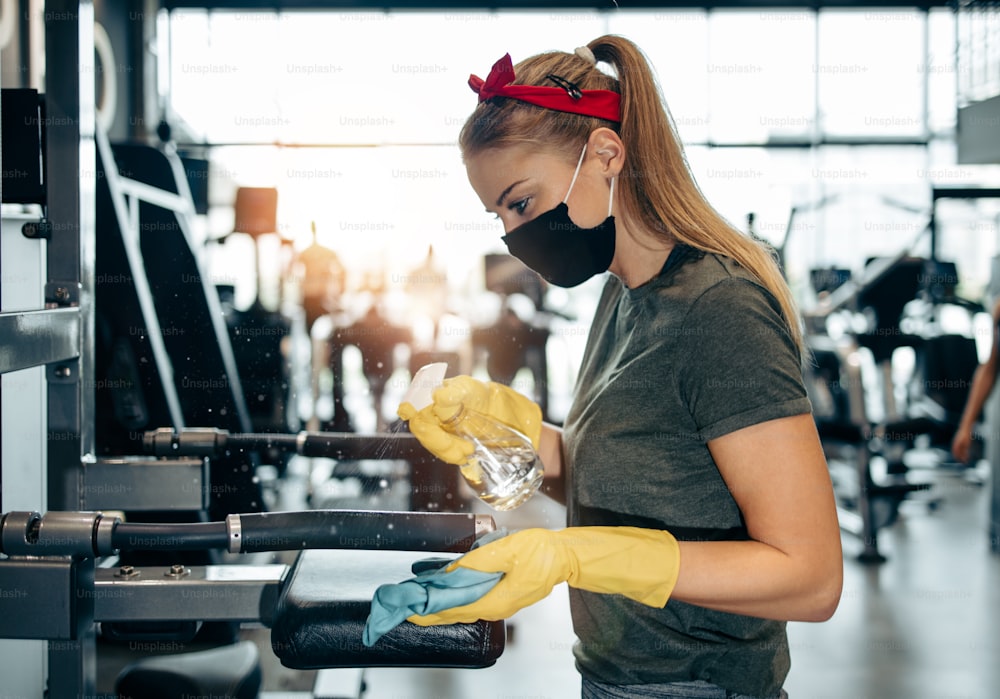 Young female worker disinfecting cleaning and weeping expensive fitness gym equipment with alcohol sprayer and cloth. Coronavirus global world pandemic and health protection safety measures.