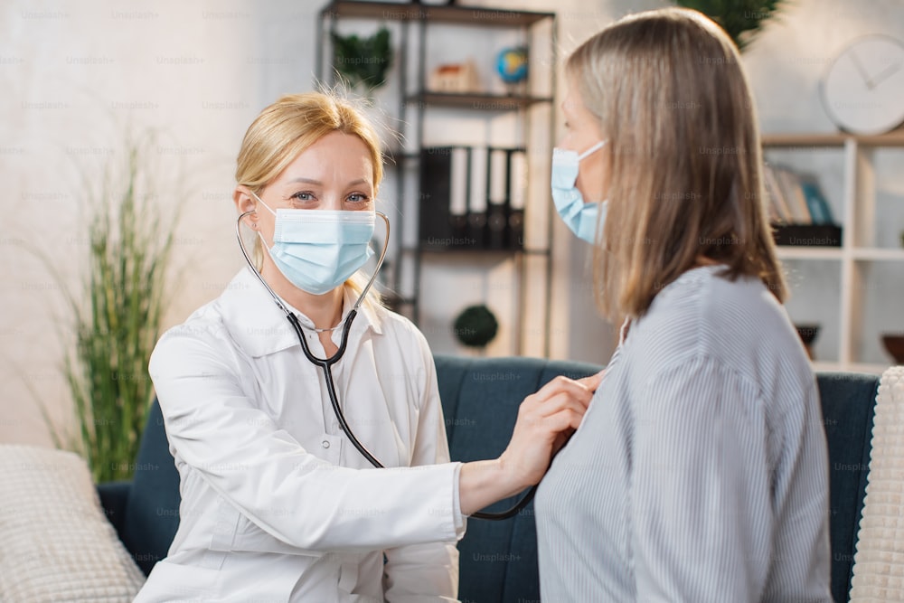 Professional experienced pleasant female doctor, wearing face mask, examining her senior woman patient and using her stethoscope for auscultation during home visit and checkup.