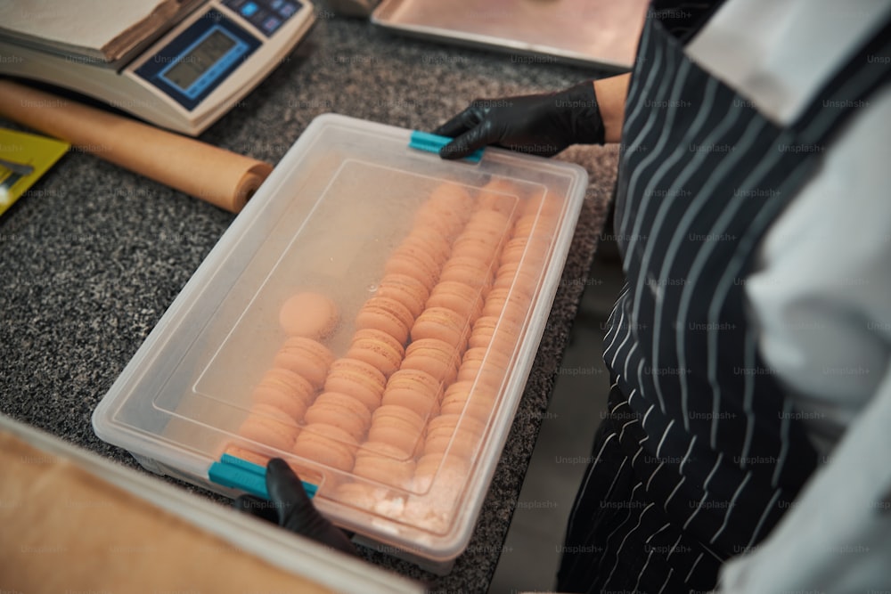 Cropped photo of chef wearing apron and gloves while holding a transparent container with lots of light-orange macarons inside
