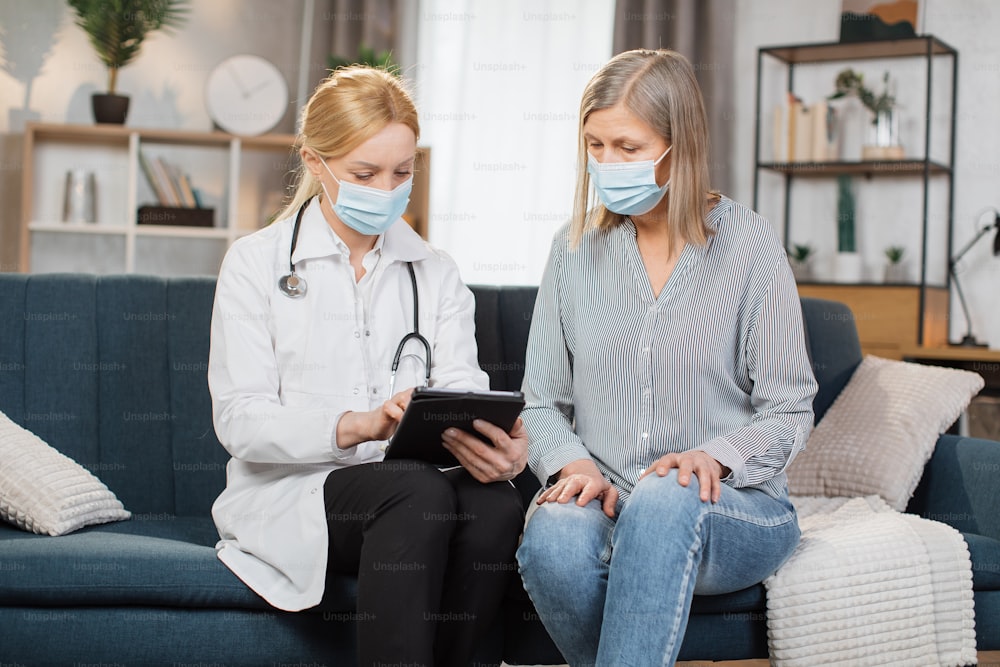 안전 보호 마스크를 착용한 의사는 COVID-19 대유행 및 독감 발생 기간 동안 가정 방문 중 노인 환자와 상담합니다. 환자와 함께 태블릿에서 결과와 약물을 보고 있는 의사