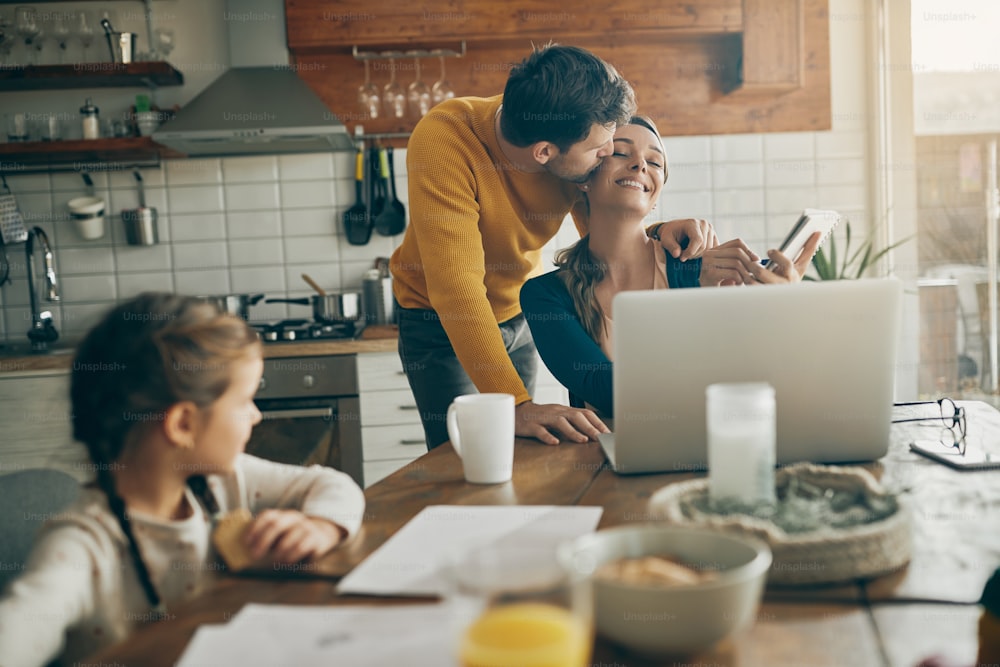 Happy woman taking notes and working on computer while husband is kissing her at home.