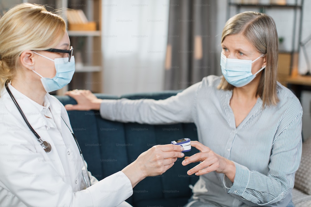 Close-up of female healthcare worker, doctor or nurse, using finger pulse oximeter, while visiting sick patient elderly woman at home, checking oxygenation level.