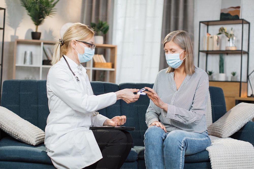 Young professional female doctor or nurse wearing protective mask, measuring saturation with pulse oximeter, while visiting her senior lady patient at home. Coronavirus covid 19 pandemic concept.