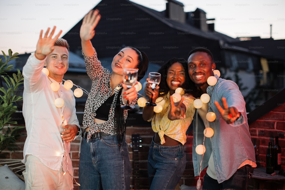 Cheerful young friends smiling and gesturing on camera during party celebration. Multicultural people in casual outfit relaxing on terrace at evening time.