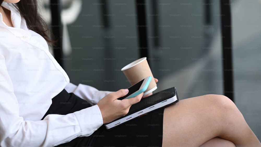 Cropped shot of female office worker holding cofee cup and using smart phone at office.