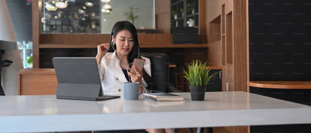Gelegenheitsgeschäftsfrau mit Kopfhörer und Blick auf das Smartphone, während sie an ihrem Arbeitsplatz sitzt.