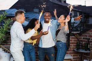 Young carefree people in casual wear having party on roof with sparklers. Four mixed race friends dancing, chatting and having fun.