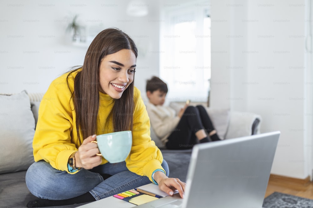 Smiling mom working at home with her child on the sofa while writing an email. Young woman working from home, while in quarantine isolation during the Covid-19 health crisis