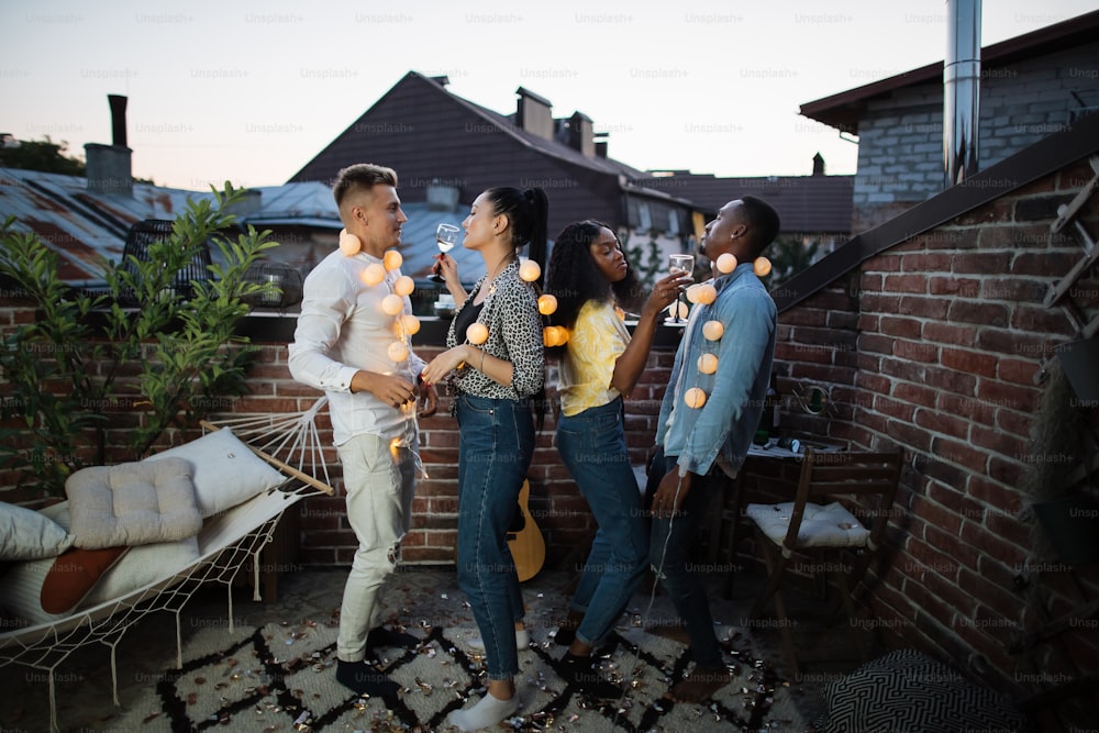 Homens e mulheres multirraciais dançando no telhado com guirlandas brilhantes no pescoço. Quatro amigos felizes em roupas casuais saindo juntos durante os fins de semana.