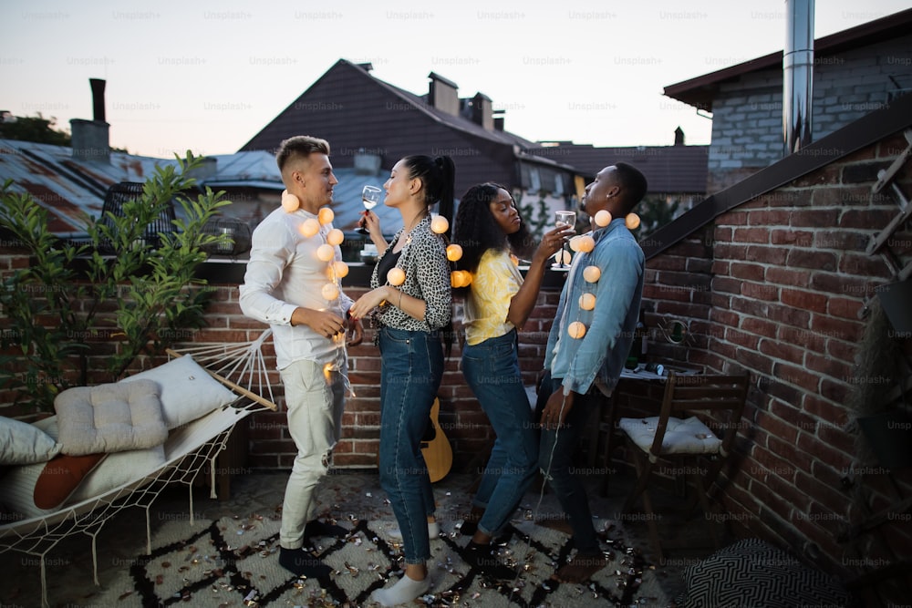 Multiracial men and women dancing on roof with glowing garlands on neck. Four happy friends in casual clothes hanging out together during weekends.